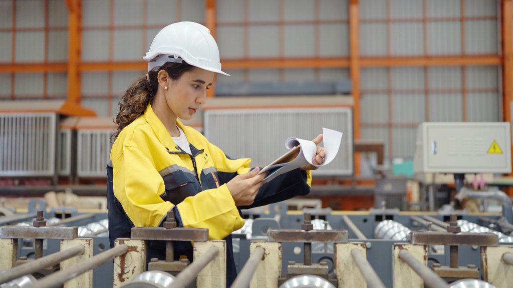Young industry worker in uniform and helmet holding clipboard checking stock of steel metal at industry manufacturing factory